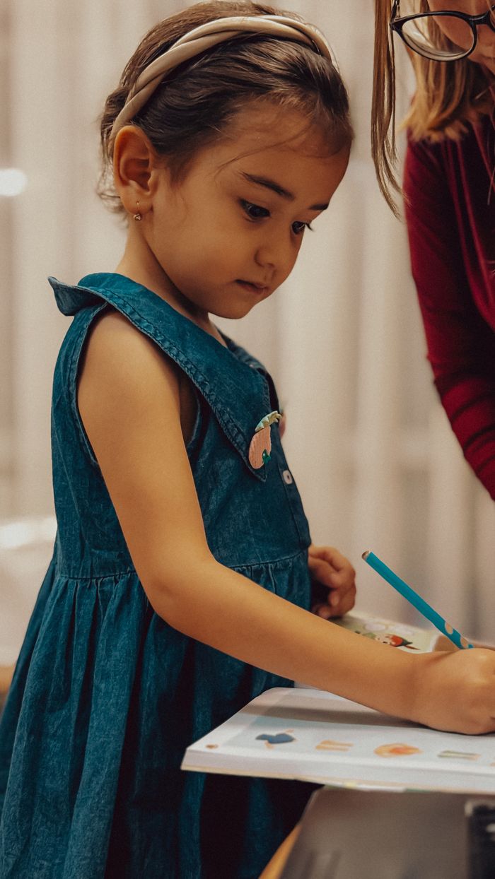 A young girl in a blue dress drawing with a pencil, focused on her artwork.